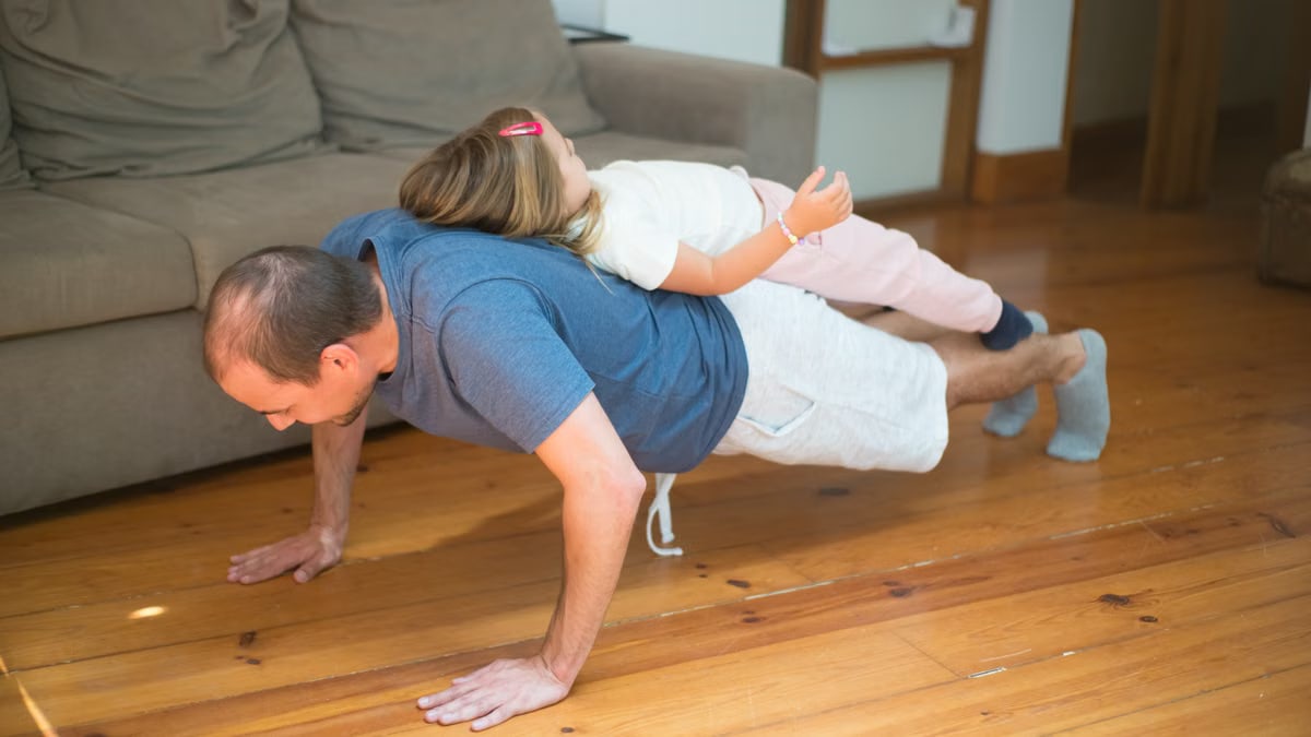 Father doing push-ups with daughter on his back in a cozy home setting.